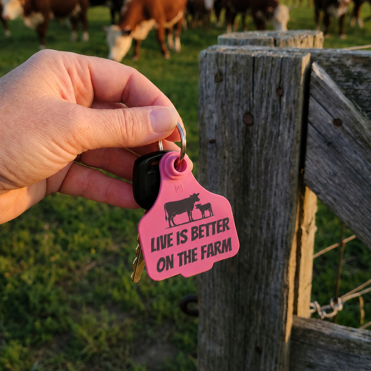 A hand holds a pink cow-shaped keychain tag with the text 'LIVE IS BETTER ON THE FARM' next to a wooden fence with grazing cows in the background.