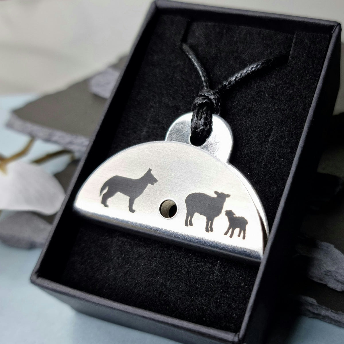 Stainless steel shepherds whistle pendant with engraved designs of a dog, sheep, and lamb in a black box.