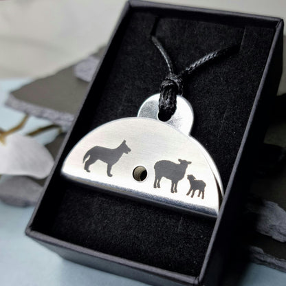 Stainless steel shepherds whistle pendant with engraved designs of a dog, sheep, and lamb in a black box.